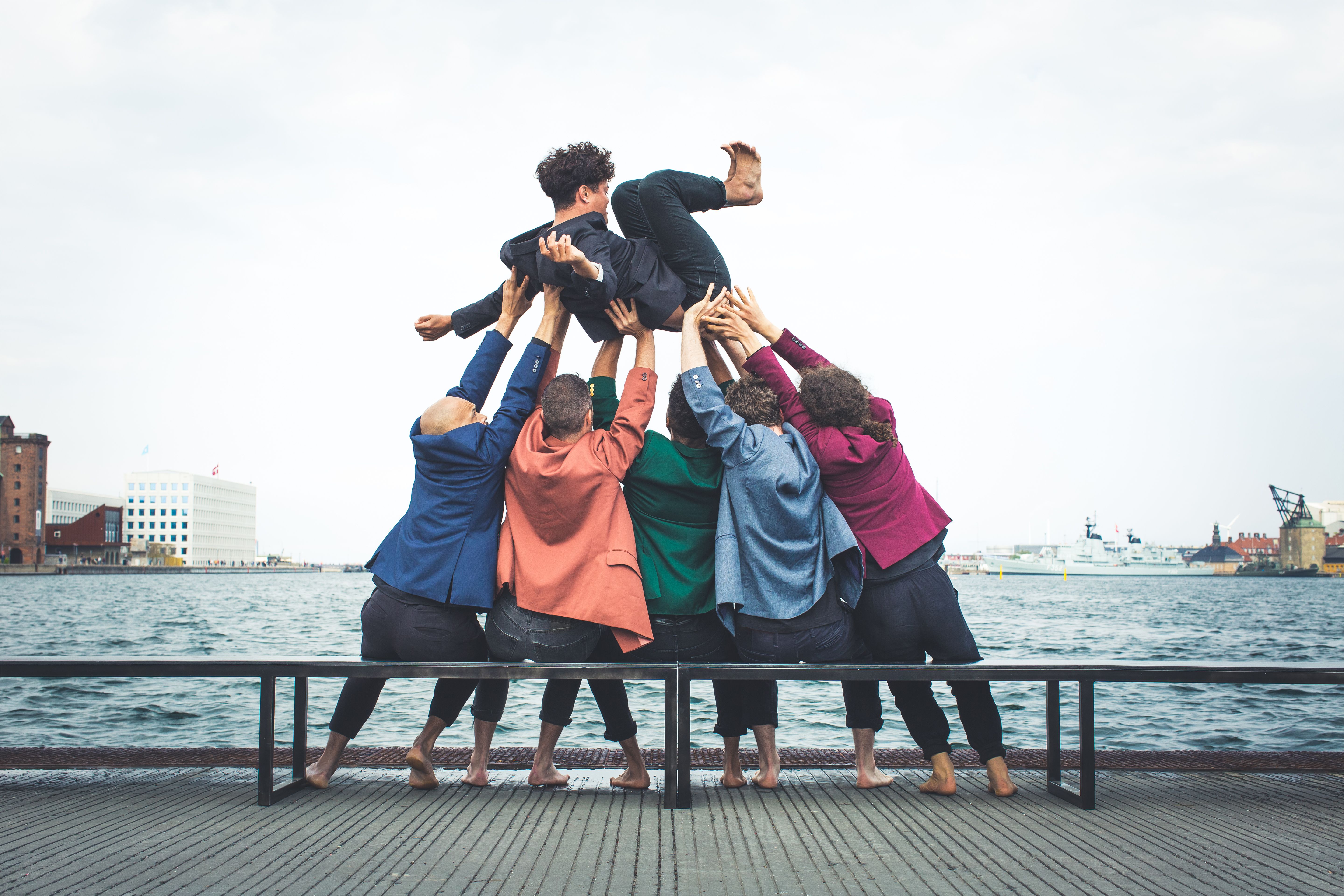 A group of seated dancers in colourful jackets holding up a dancer above their heads