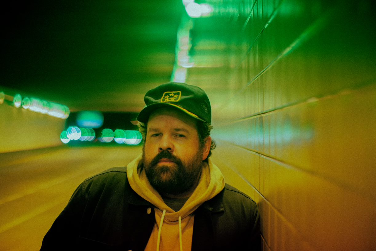 Donovan Woods wearing a hoodie and ballcap standing in front of a yellow and green background