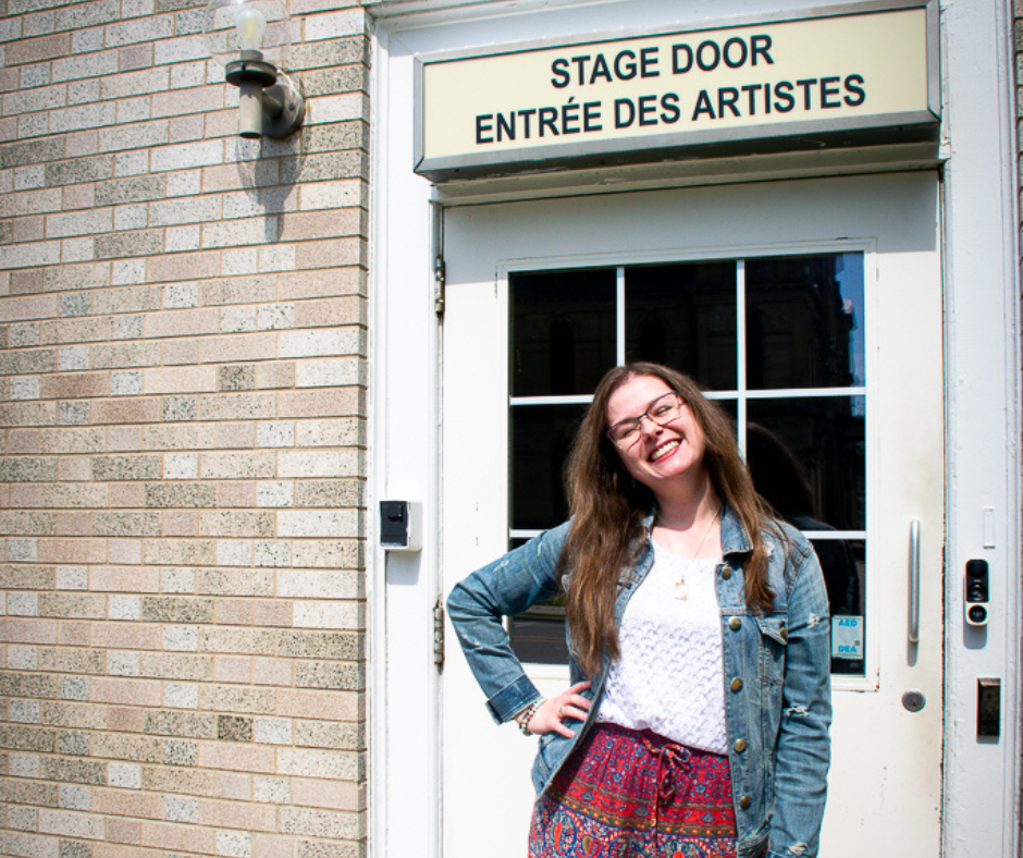 Katelyn Goodwin stands in front of the stage door entrance of the Fredericton Playhouse. Her long brown hair is being hit by the sun, she stands smiling while wearing a jean jacket and colourful red pants.