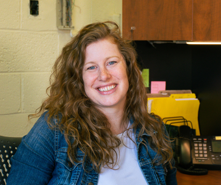 Meghan Callaghan, curly red hair, sits at her desk smiling. She is wearing a denim jacket and a white t-shirt.