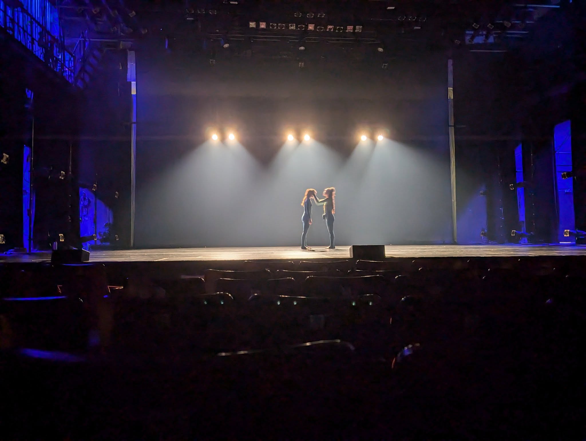 A wide shot of the stage from the audience's perspectve. With a cool dark blue lighting, two dancers stand centre stage and caress each other.