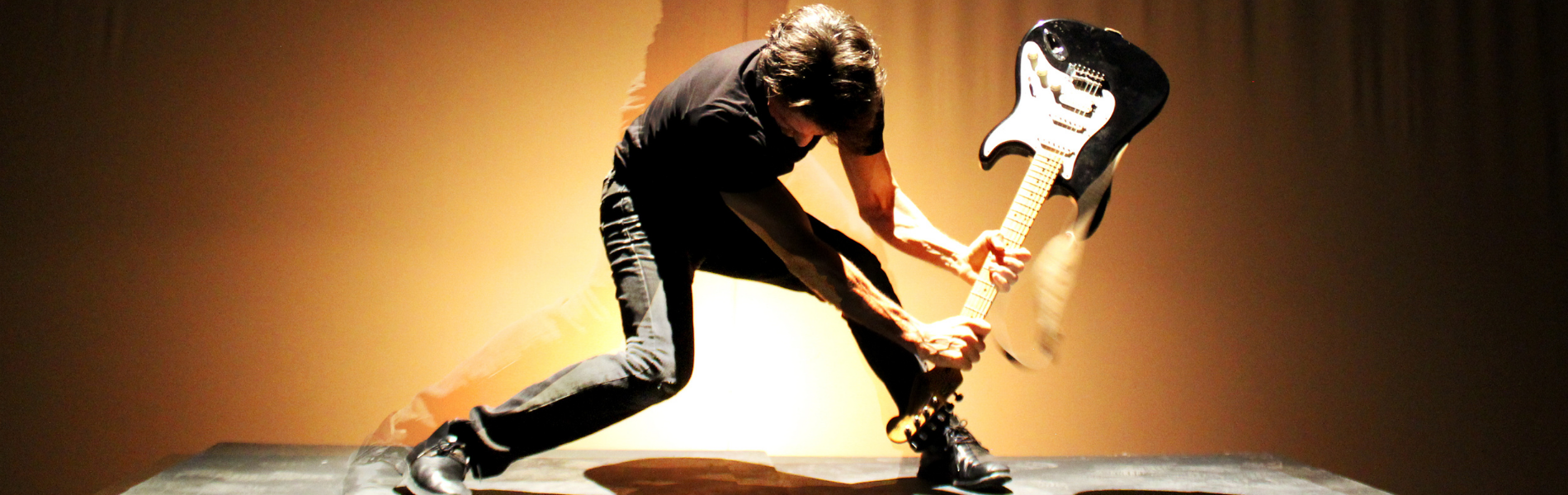 A person in a black tshirt bent over about to smash an electric guitar on the ground
