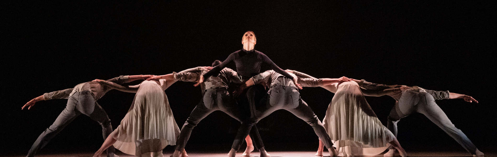 A group of dancers from Ballet Kelowna: one in the centre is standing and looking up at the ceiling, the others are bent over with their backs to the camera, arms outstretched