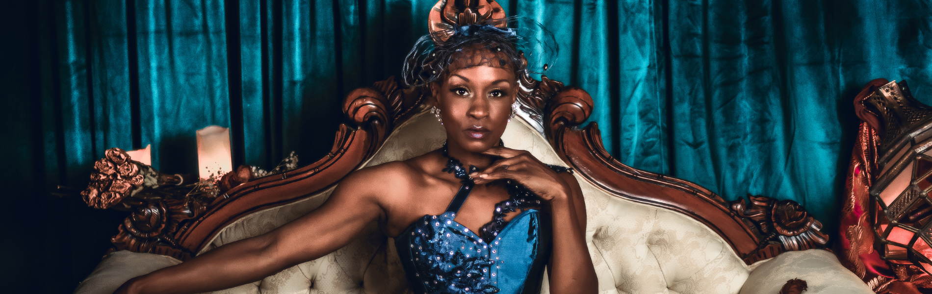 A close up of Tymisha Harris wearing a royal blue strapless top with a fascinator on her head. She's sitting on a regal looking cream couch with wooden details, looking straight on to the camera with no smile