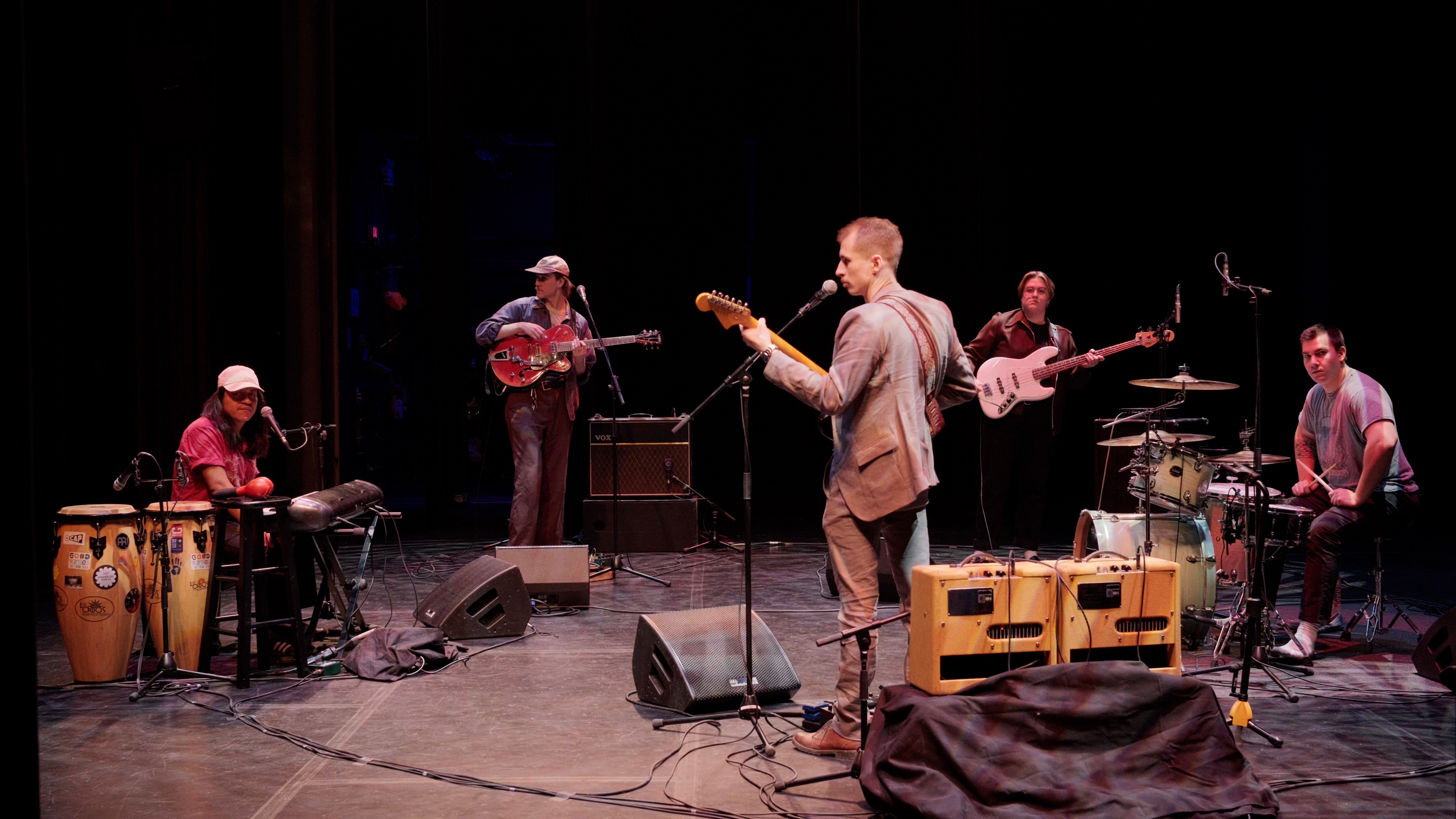 Five members of the band circle up and play music together. There are drums, guitars, and a keyboard.