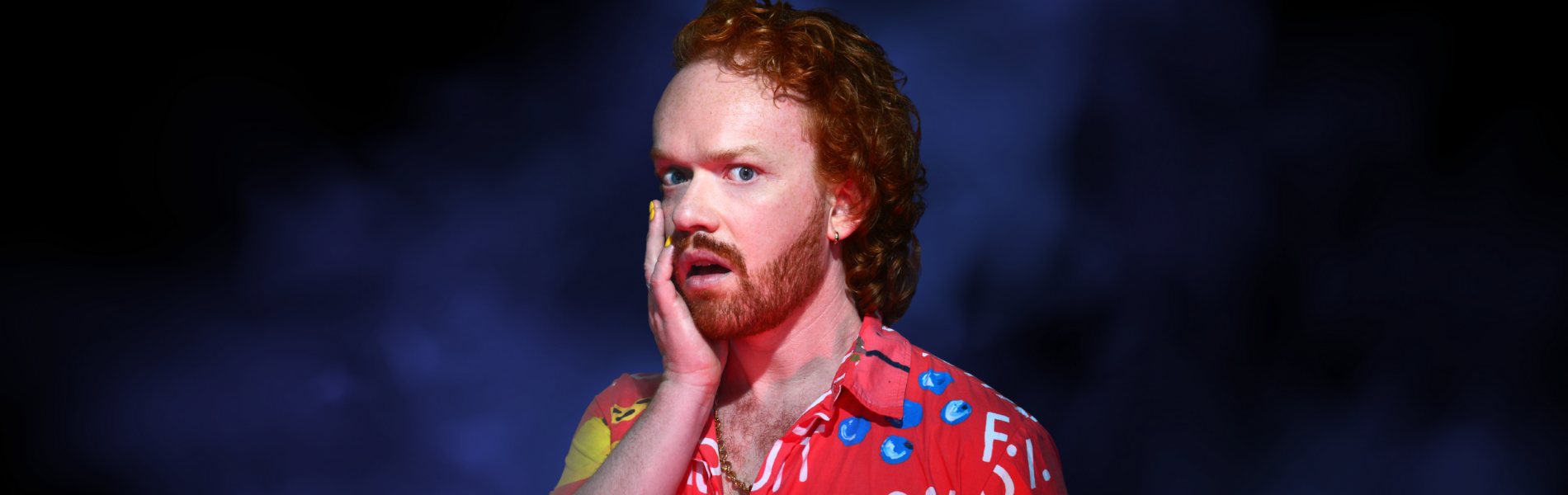 Performer Johnnie McNamara Walker against a dark background, wearing a red collared shirt holding his hand to his face, looking shocked