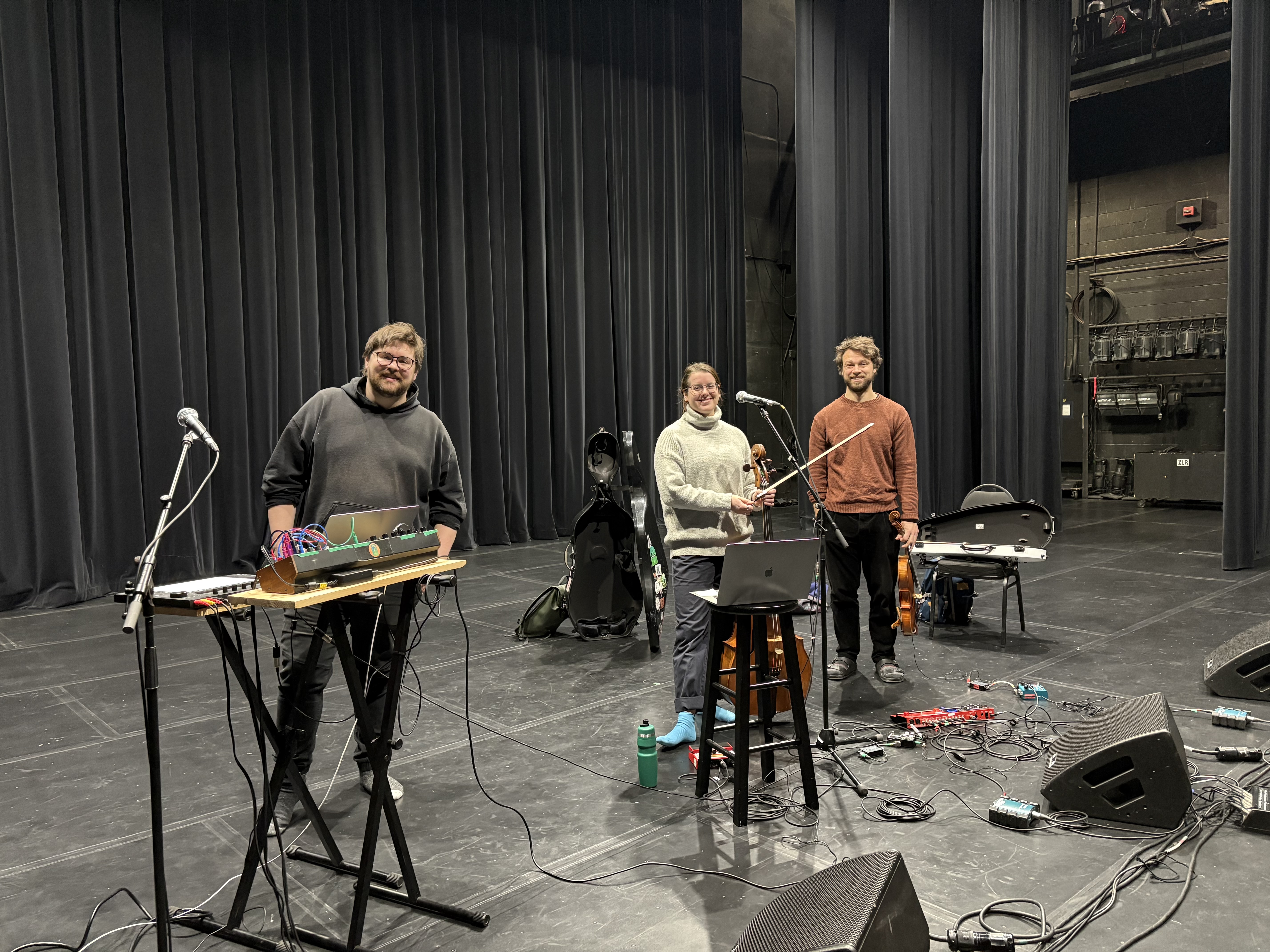 Three band members of Palmer smile and face the camera. On stage around them are various instruments and pieces of recording equipment.