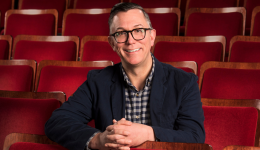 Jeff Richardson in front of red theatre seats wearing a checkered shirt and dark blue jacket