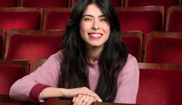 Lulu sits in the Playhouse theatre in a pink sweater, enthusiastically smiling at the camera.
