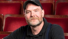 Greg sits in the Playhouse audience, arms crossed, all dressed in black.