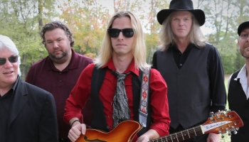 A group of men standing in a field. One is holding a guitar.