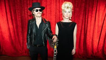 Two people dressed in black standing against a red curtain background