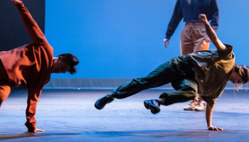 Two street dancers against a blue background, balanced sideways on their arms