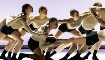 a group of dancers in light skin toned tops, black shorts and socks all leaning together as a group while once dancer is held horizontally in front