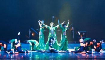 a group of performers crouched on a stage with a dark blue background and green lighting, three performers stand in the middle with their arms extended