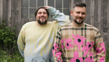 An image of Darcy & Jer standing in front of a graying fence with greenery around.