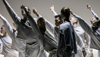 A shaded group of dancers dressed in white against a white background, arms extended up to the left