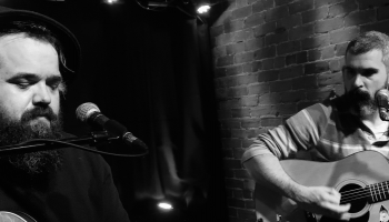A black and white image of Stephen Lewis and Bobby Mahar playing guitar and singing on stage.