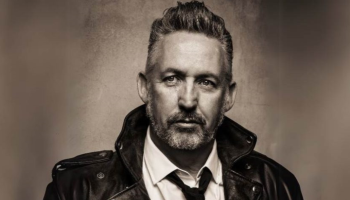 A sepia toned image of Harland Williams in a leather coat and tie.