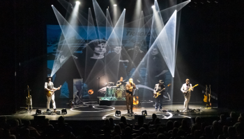 A rock band set up on stage with white spotlights on the band. An image of the Tragically Hip projected on a screen behind the band. 