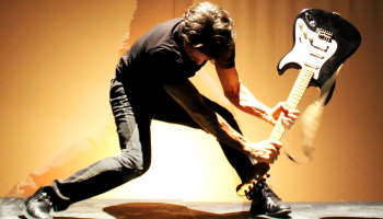 A person in a black tshirt bent over about to smash an electric guitar on the ground