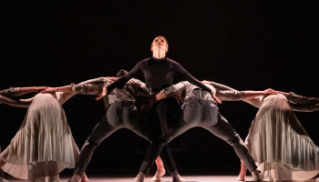 A group of dancers from Ballet Kelowna: one in the centre is standing and looking up at the ceiling, the others are bent over with their backs to the camera, arms outstretched