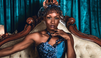 A close up of Tymisha Harris wearing a royal blue strapless top with a fascinator on her head. She's sitting on a regal looking cream couch with wooden details, looking straight on to the camera with no smile
