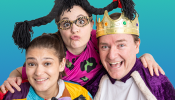 Three performers of DuffleBag Theatre huddled together in the centre of the photo, each are smiling and dressed in capes and costume pieces
