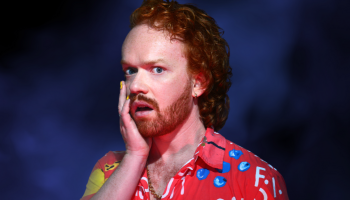 Performer Johnnie McNamara Walker against a dark background, wearing a red collared shirt holding his hand to his face, looking shocked