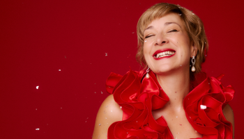 Jill Barber smiling as she looks away from the camera, she's wearing a red sparkly dress for the holidays, photographed against a red background