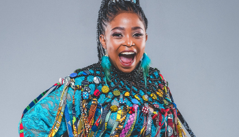A close up of Nomfusi in a light blue tassled outfit, pictured from the chest up. She is looking right at the camera with her mouth open in a big, energetic smile