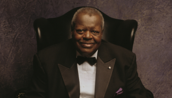 A closely cropped image of Oscar Peterson from the waist up, sitting in a high backed chair wearing a black tuxedo jacket, white shirt, and black botwie