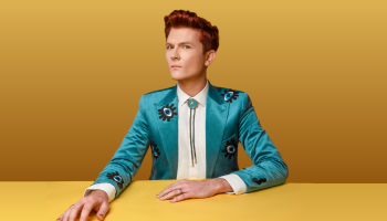 Rhys Nicholson sitting at a yellow table surface against a darker yellow background. They are wearing a light teal blue jacket and a white shirt, looking at the camera with their head turned slightly sideways