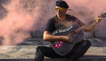 Tom Morello sitting on a sidewalk holding a black acoustic guitar in his lap. He's wearing reflective sunglasses, looking away from the camera, with pink smoke surrounding him