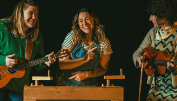 Three performers of Tunes and Tall Tales standing in front of a black background, each holding an instrument and smiling at each other