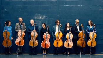 The members of the band hold Cellos, standing against a wall.