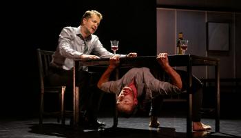 A dancer is upside down, holding onto the bottom of a table