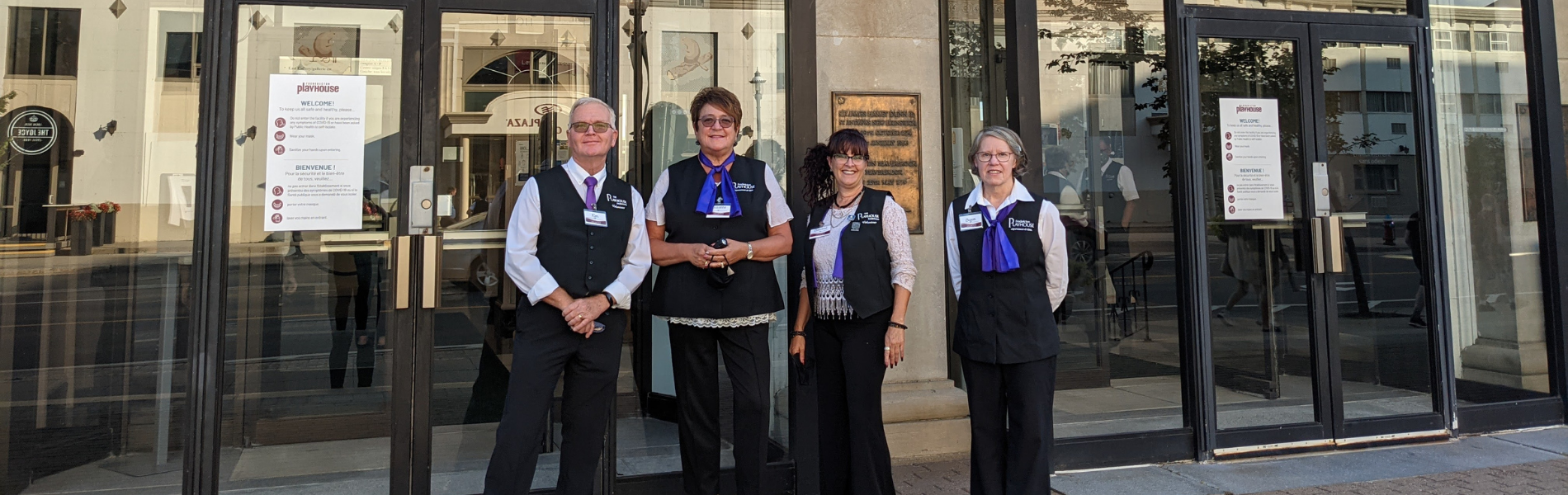 A man and three women in black Playhouse volunteer vests and purple scarves outside the Playhouse front doors