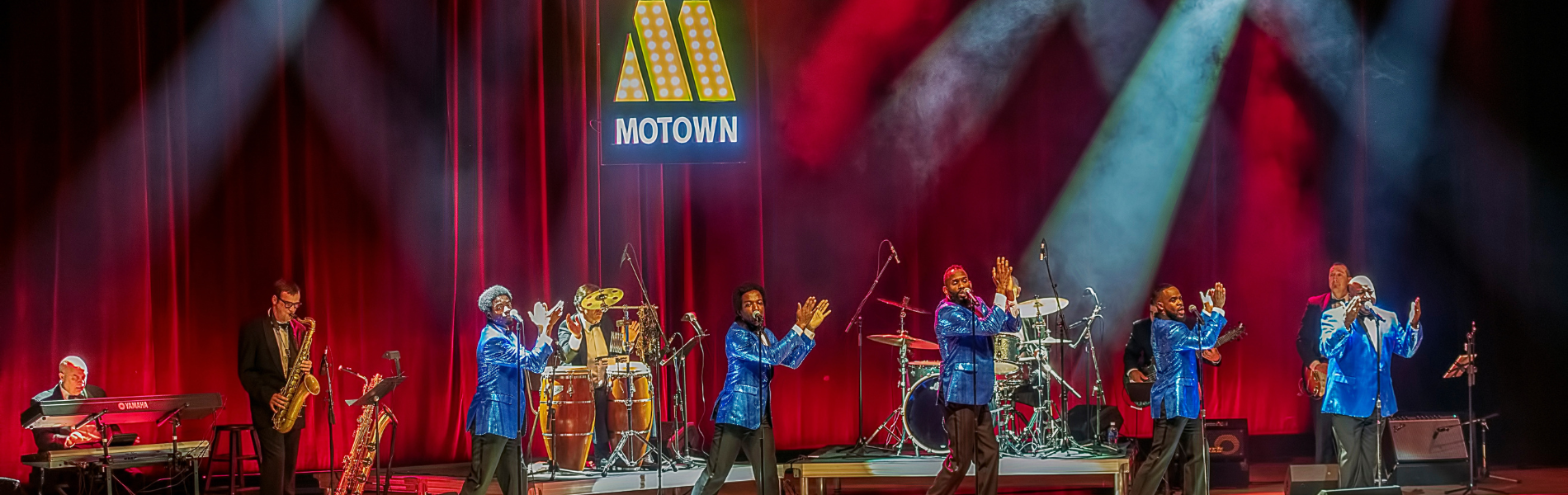 5 men in blue suit coats dancing in sync with musicians behind them and a sign that says Motown.