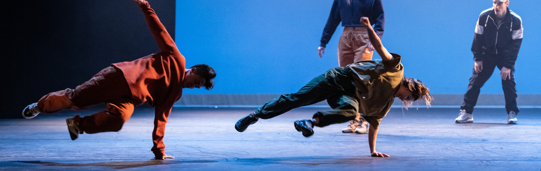 Two street dancers against a blue background, balanced sideways on their arms