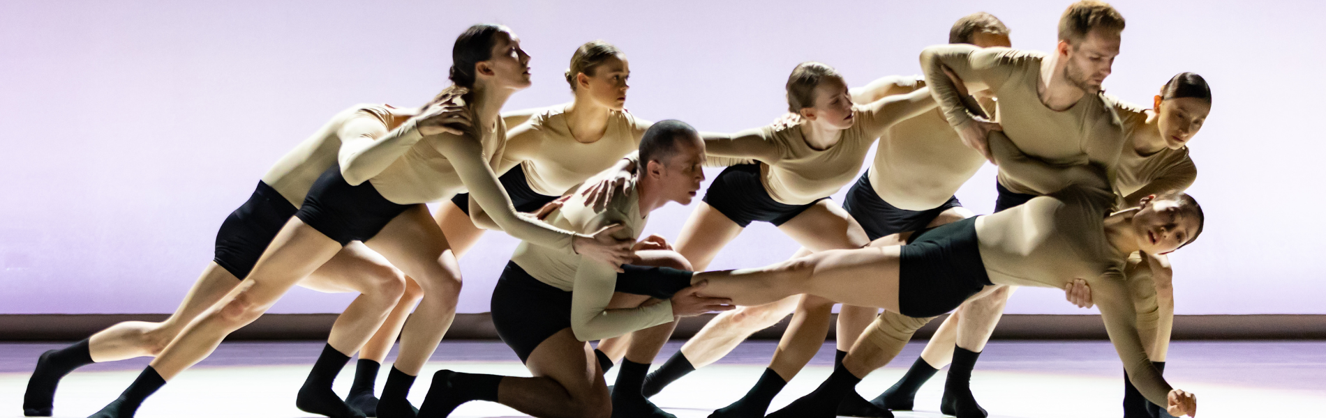 a group of dancers in light skin toned tops, black shorts and socks all leaning together as a group while once dancer is held horizontally in front