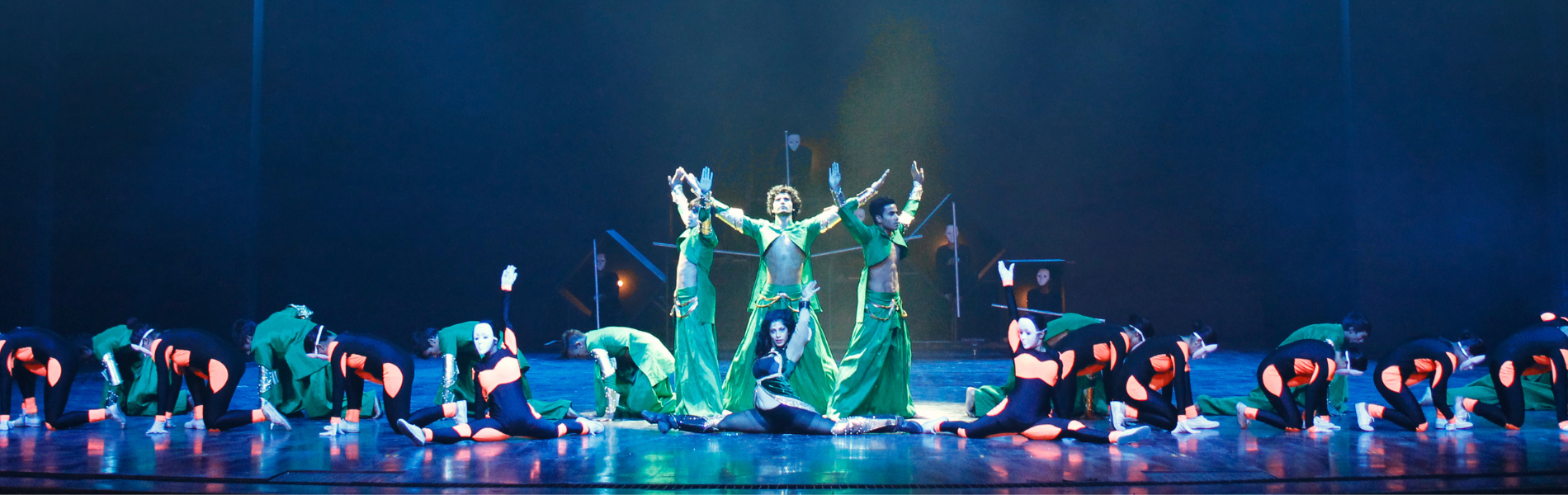 a group of performers crouched on a stage with a dark blue background and green lighting, three performers stand in the middle with their arms extended