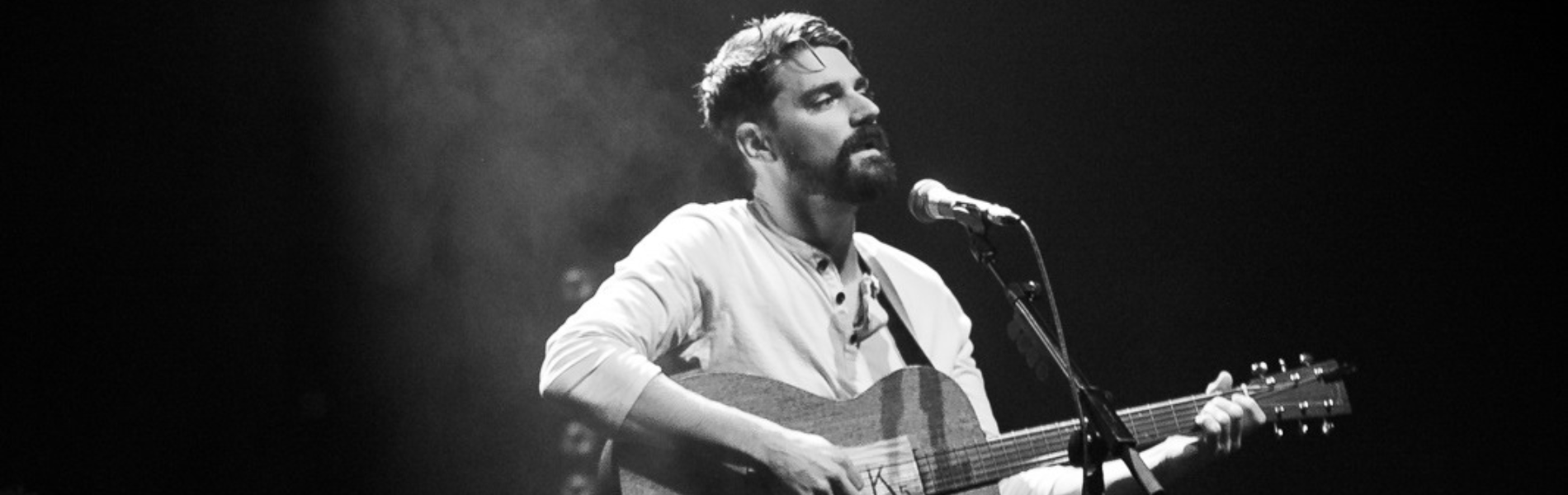 A black and white image of Tim Baker in the spotlight, singing and playing guitar.