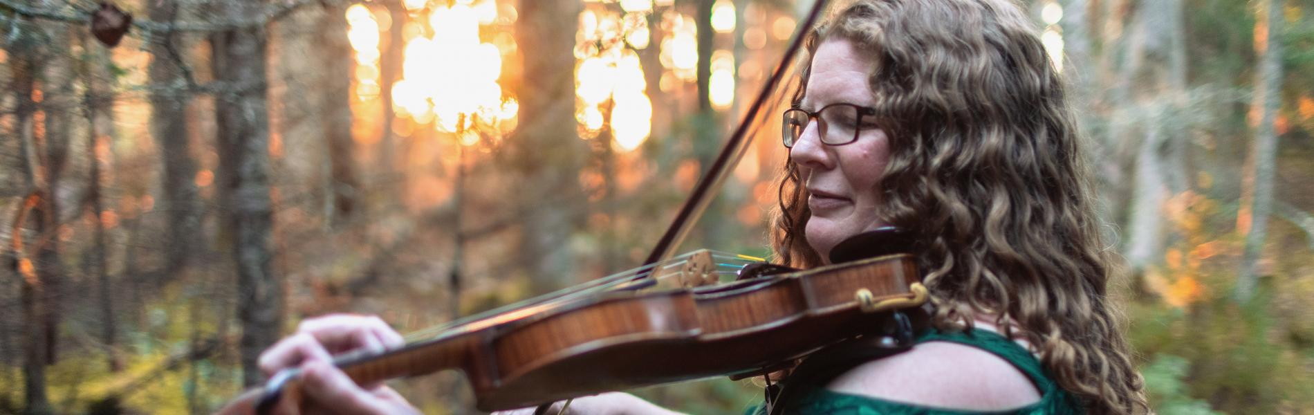 Katherine Moller playing the fiddle outside with trees and a sunset in the background.