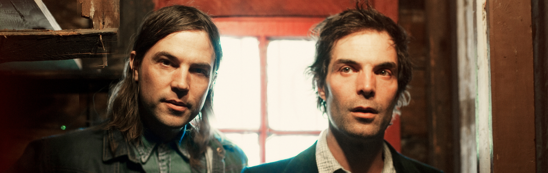A close up of the two Barr Brothers standing in front of a window looking to the side off-camera 