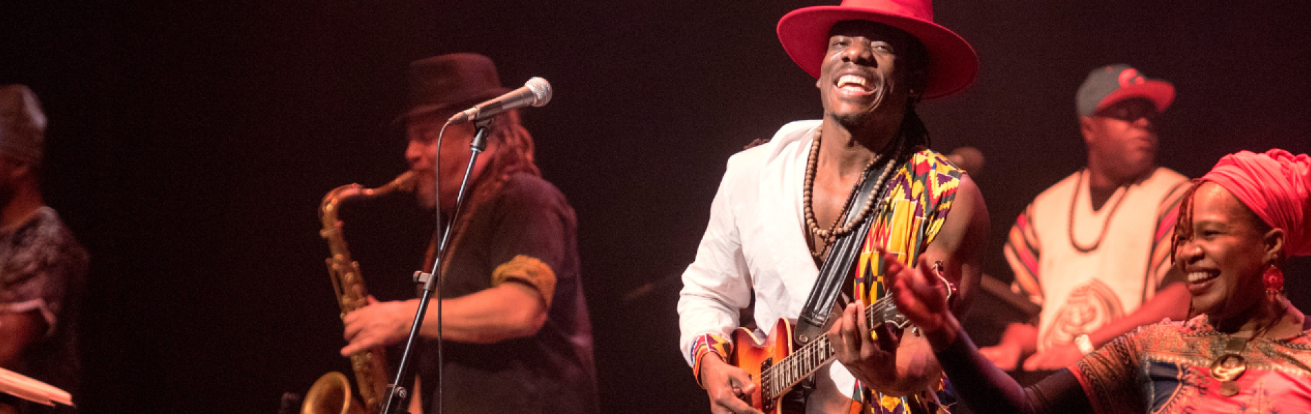 A close up image of 5 artists smiling and playing instruments on stage, the whole image has a dark background and warm lighting. 