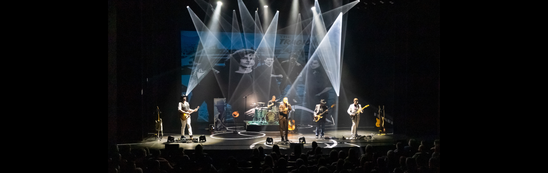 A rock band set up on stage with white spotlights on the band. An image of the Tragically Hip projected on a screen behind the band. 