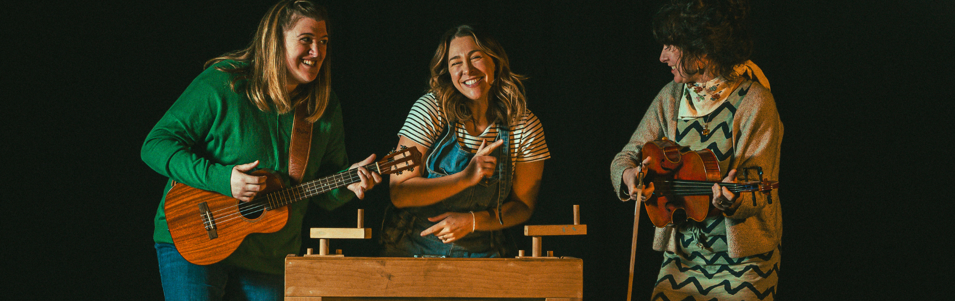 Three performers of Tunes and Tall Tales standing in front of a black background, each holding an instrument and smiling at each other