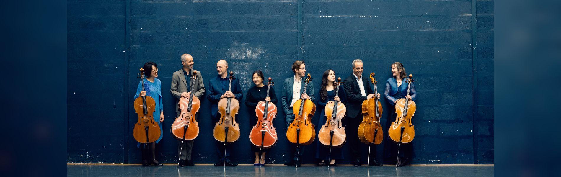 The members of the band hold Cellos, standing against a wall.