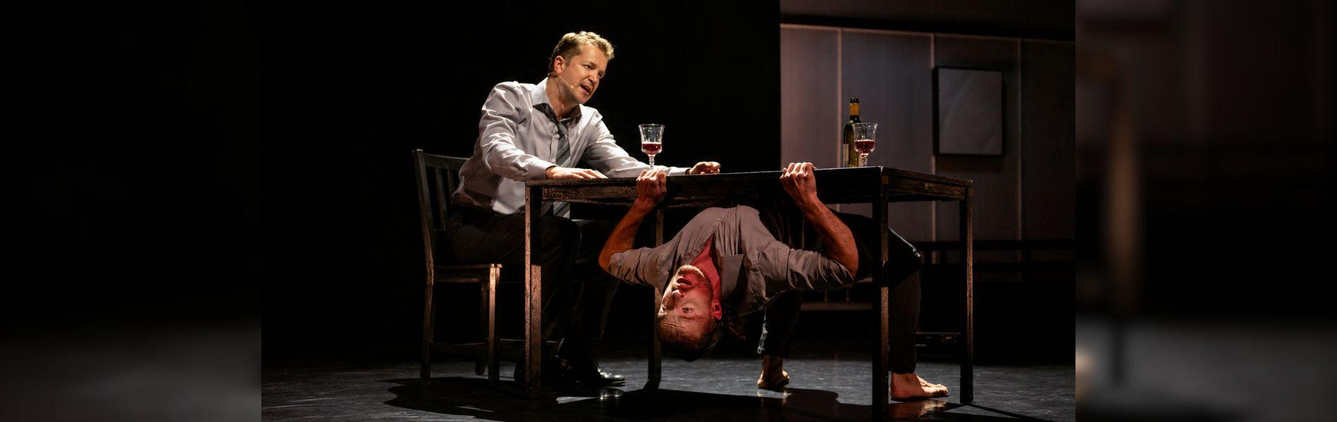 A dancer is upside down, holding onto the bottom of a table